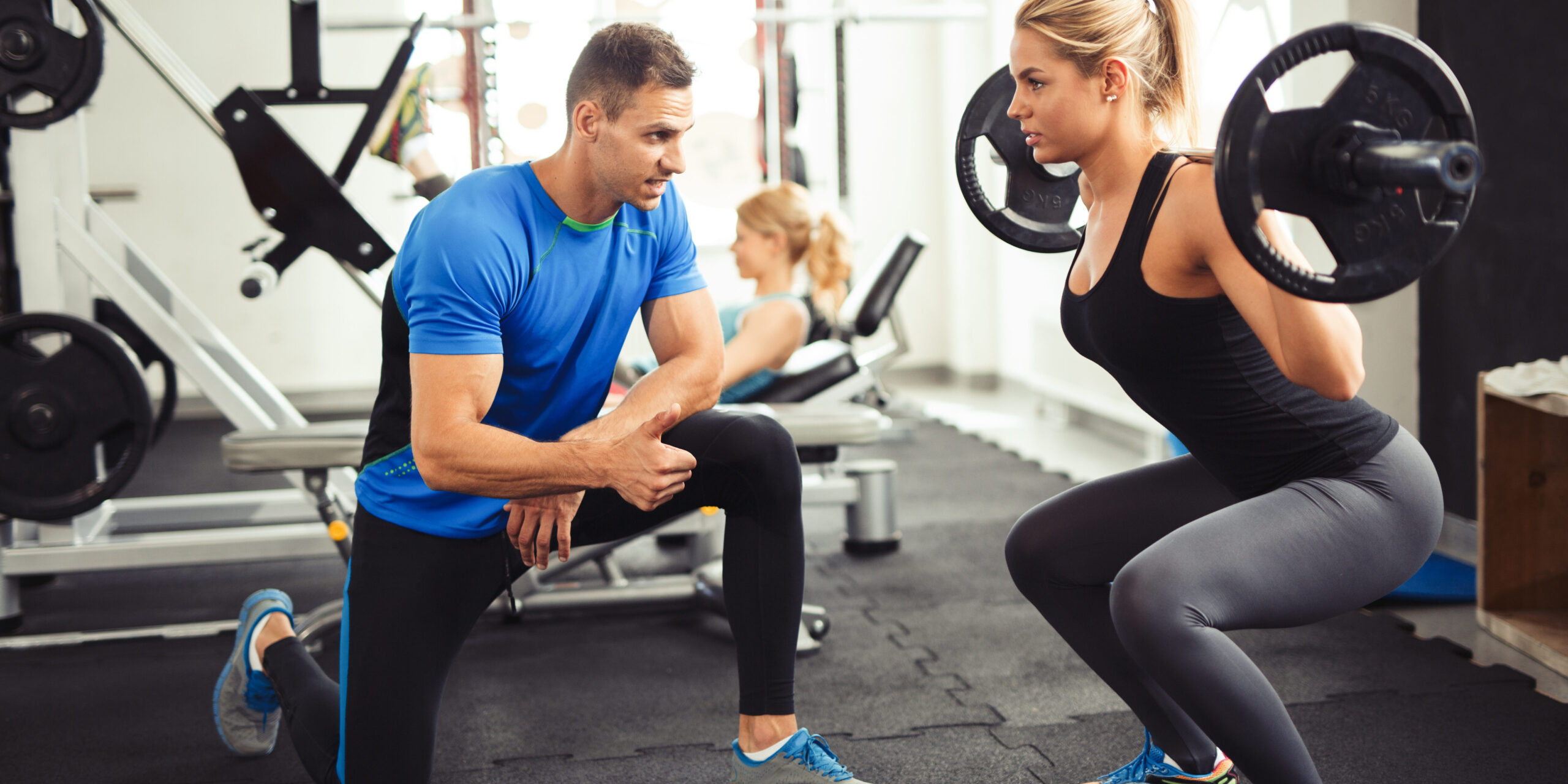 Young woman lifting barbell with her personal trainer motivation.